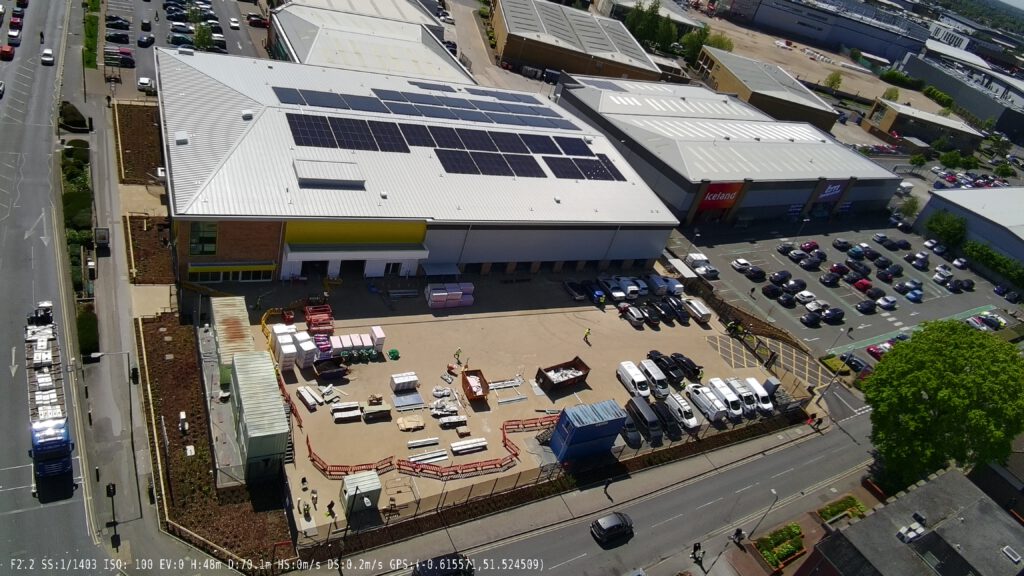 single Photo Aerial view of a Big Yellow Self Storage facility. Large building with bright yellow and blue sections, white roofs covered in solar panels. Parking area in front with a few vehicles. Surrounded by roads and green spaces. Shows modern commercial architecture with sustainable energy features.